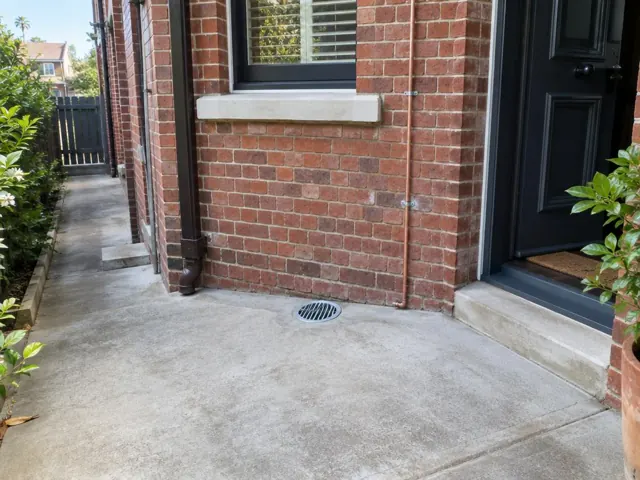 Outdoor concrete gully grate beside a brick house and side path, with a dark front door in Double Bay