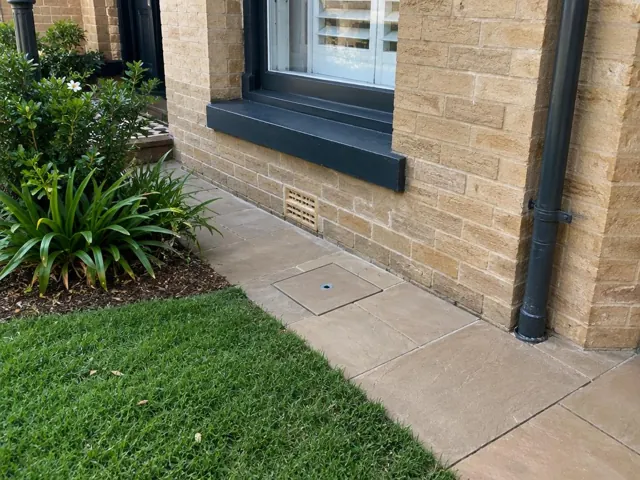 Flush access cover set in paved path beside a brick house and garden in Paddington