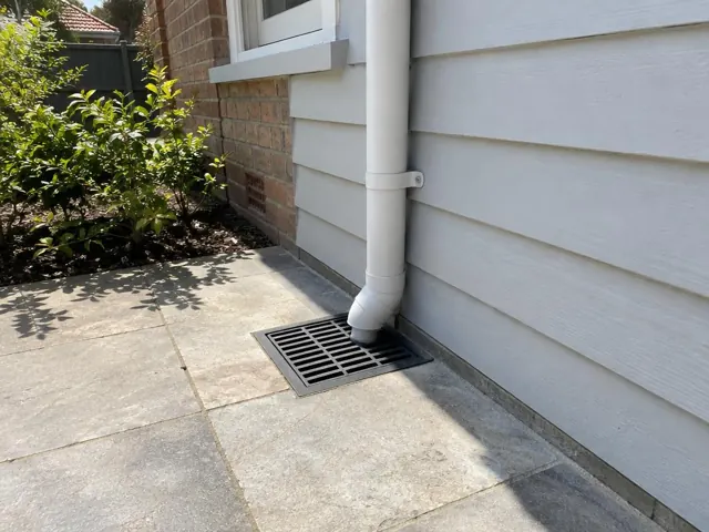Stormwater grate beside a white weatherboard house on a paved side path in Kensington